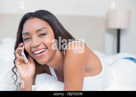 Afroamerikanische Frau, die ihr Smartphone hält, während sie sich auf dem Bett mit einer Lampe auf dem Nachttisch im Schlafzimmer zurücklehnt Stockfoto