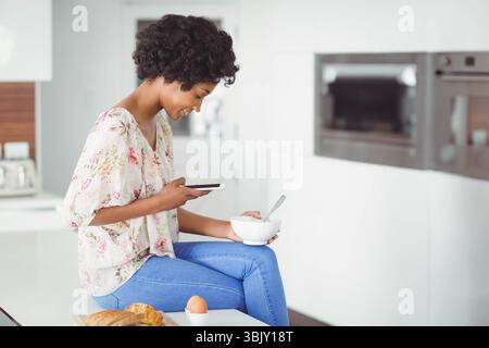 Afroamerikanerin sitzt auf der Küchenarbeitsplatte und hält Smartphone und Schüssel in der Küche zu Hause Stockfoto