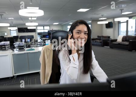 Frau in weißer Bluse mit Headset-Ohrhörer, lächelt und arbeitet am Schreibtisch Stockfoto