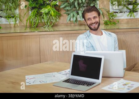 Mann, der an einem Holztisch in einem modernen Arbeitsbereich sitzt und Designskizzen und Laptops überprüft, Kopierraum Stockfoto