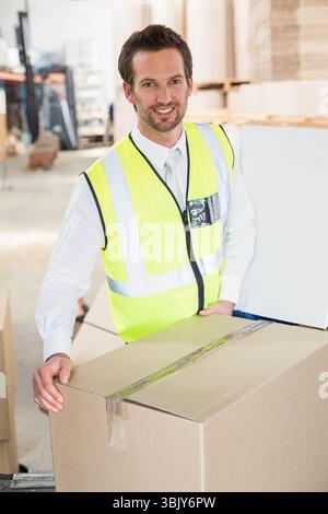 Mann mit neongelber Sicherheitsweste, der versiegelte Pappschachtel im Lager mit Metallregalen hält Stockfoto