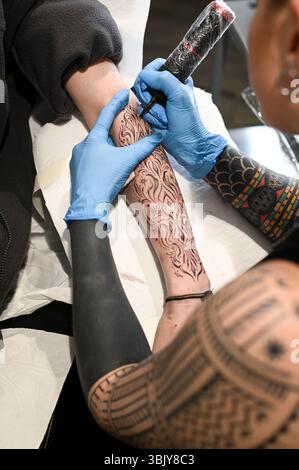 Tätowierer trägt blaue Handschuhe und zeichnet ein Tattoo mit schwarzer Tinte auf den Arm des Klienten in einem Tätowierstudio Stockfoto