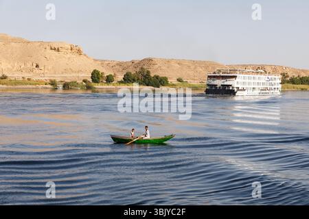 Zwei Personen rudern ein kleines grünes Boot auf einem Fluss, mit einem großen Kreuzfahrtschiff im Hintergrund. Stockfoto