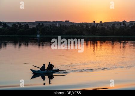 Zwei Männer rudern bei Sonnenuntergang auf einem Fluss, wahrscheinlich für Freizeit oder Transport, mit Sonnenuntergang hinter einer fernen Küste. Stockfoto