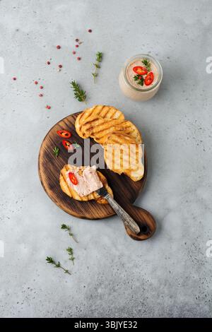 Hausgemachte Hühnerpastete aus Leber mit rotem Pfeffer, Thymianzweigen und gerösteten Brotscheiben auf altem Holzbrett auf grauem Beton oder s Stockfoto