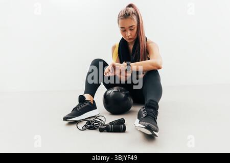 Junge Frau in Sportkleidung mit einem Handtuch am Hals sitzt drinnen mit Blick auf Smartwatch am Handgelenk Stockfoto