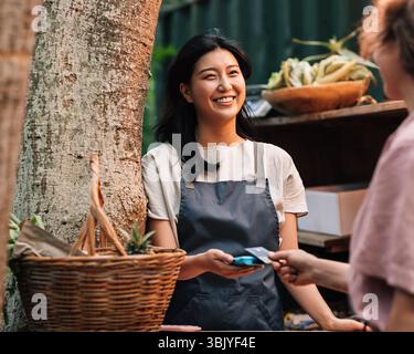Glückliche asiatische Frau auf einer Schürze, die auf dem Markt bezahlt wird. Anbieter mit einem Kartenautomat, der lächelt und den CUS ansieht Stockfoto