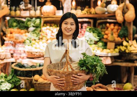 Lächelnde asiatische Frau, die einen Korb mit Gemüse hält. Junge Frauen, die draußen auf dem Bauernmarkt stehen. Stockfoto