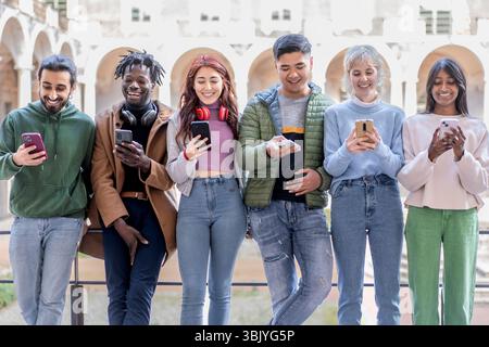 Lächelnde multikulturelle Gruppe junger Menschen, die auf Smartphones schauen, während sie zusammen in einer städtischen Umgebung stehen und digitale Interaktion genießen Stockfoto