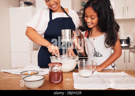 Seniorin und Enkelin, die Mehl an einem Tisch in der Küche sieben. Das Mädchen und ihre Oma machen Teig. Stockfoto