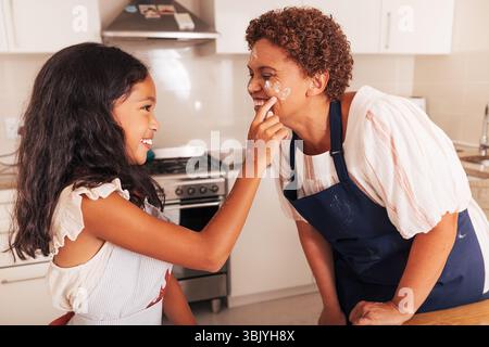 Die Enkelin berührte die Wange ihrer Großmutter mit einem mit Mehl bedeckten Finger. Glückliches Mädchen und Großmutter, die Spaß in der Küche haben. Stockfoto