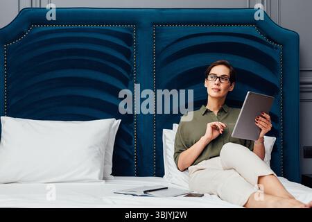 Junge Frau hält ein digitales Tablet und schaut weg, während Liegen auf einem Bett in einem Luxus-Hotelzimmer Stockfoto