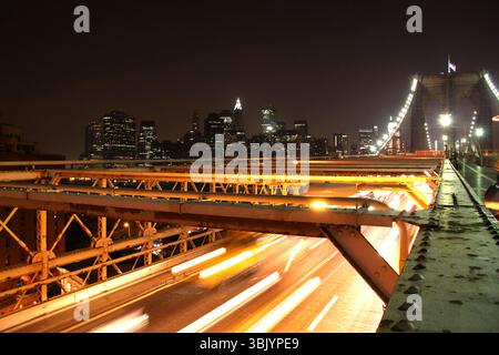 Lange Zeit ist der Verkehr auf der Brooklyn Bridge nachts ausgesetzt, mit der Skyline von Manhattan im Hintergrund und Lichtspuren über die Straße. Stockfoto