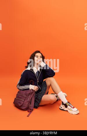 Eine junge Frau im trendigen Outfit sitzt anmutig vor einem orangen Hintergrund und strahlt Selbstvertrauen aus. Stockfoto