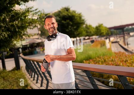 Ein fröhlicher Mann steht am Wasser, sonnt sich im Sonnenlicht mit einem warmen Getränk in der Hand, Kopfhörern um den Hals und genießt die ruhige Atmosphäre. Stockfoto