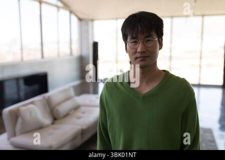 Asiatischer Mann mit grünem Pullover und Brille, der in der Lounge mit hohen Fenstern neben einem Ledersofa steht Stockfoto