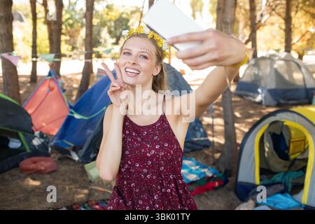Frau mit floralem Stirnband und Selfie mit Smartphone im Waldcamp mit Zelten und Fahnen Stockfoto