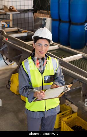 Inspektorin inspiziert Förderband in der Anlage mit Helm Ohrenschützern Brille mit Klemmbrett Stockfoto