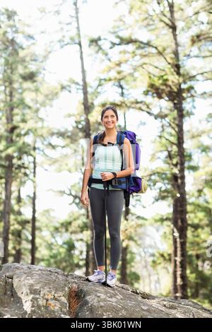 Weibliche Wanderer, die auf Felsen in Kiefernwäldern steht und Stöcke mit violettem Rucksack mit Polster trägt Stockfoto