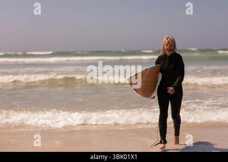 Weibliche Surferin in schwarzem Neoprenanzug, die aus der Brandung am Sandstrand mit Holzkorn-Shortboard auftaucht Stockfoto
