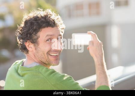 Mann mit grünem Pullover, der auf einem Balkongeländer steht und ein Telefon für Selfie mit verschwommenen Fassaden hält Stockfoto
