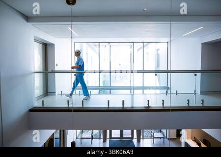 Asiatische Frau in Schutten, die entlang des Krankenhausflugs mit Glasgeländer und Fenstern, Kopierraum läuft Stockfoto