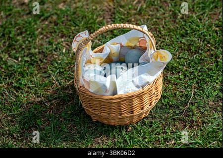 Ein gewebter Korb enthält farbenfrohe dekorative Eier, die draußen auf einem Stoffbett liegen. Stockfoto