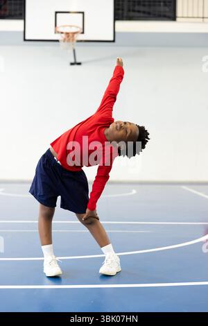 Der afroamerikanische Junge dehnt sich fröhlich in der Turnhalle der Schule, trägt ein rotes Hemd und blaue Shorts Stockfoto