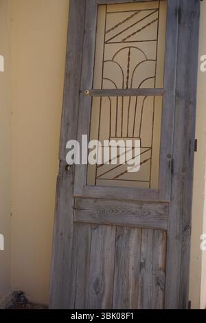 Eine alte, dekorative Holztür, die an eine blassgelbe Wand gelehnt ist. Die Tür verfügt über eine Glasscheibe mit einem komplexen geometrischen und linearen Design Stockfoto