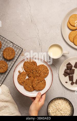Oben auf eine Oberfläche mit einem Keksständer, Hafer, geschredderter Schokolade und einer Tasse Hafermilch. Eine Hand legt eine Schüssel mit Keksen neben sie. Stockfoto