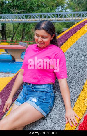 Halbkörperporträt einer Latina in einem pinkfarbenen Oberteil und einer Jeansshorts, die mit einem Bein steht, während sie in einem entspannten, nachdenklichen Look nach unten blickt Stockfoto