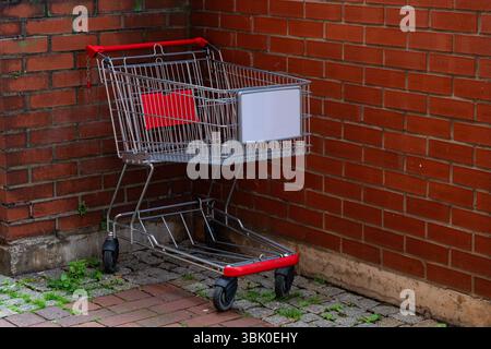 Ein Einkaufswagen steht in einer Ecke leer und lehnt sich an eine verwitterte Ziegelwand. Die roten Akzente auf dem Wagen stehen im Kontrast zu den Erdtönen des Surro Stockfoto