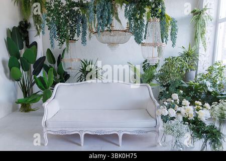 Elegantes Wohnzimmer mit weißem Sofa, üppigen Zimmerpflanzen, Kristallleuchtern und Blumenarrangements in stilvoller botanischer Einrichtung. Stockfoto