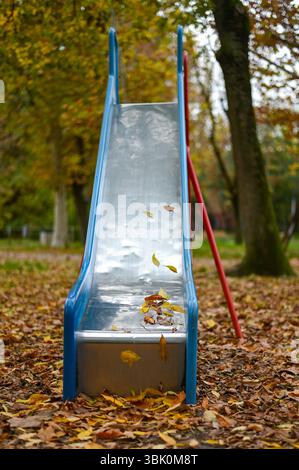 Leere Kinderrutsche mit Herbstlaub bedeckt in einem einsamen Park, umgeben von Bäumen mit Herbstlaub. Stockfoto
