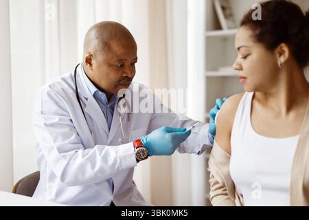Schwarzer Doktor, der Handschuhe trägt, um eine junge Patientin zu impfen Stockfoto