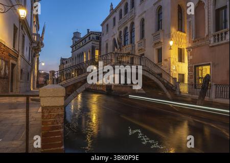 Kleine Fußgängerbrücke, die sich in der Dämmerung über den Kanal in venedig, italien, mit Lichtspuren, die sich vom vorbeifahrenden Boot im Wasser spiegeln Stockfoto