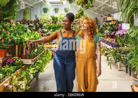 Zwei junge Frauen gehen durch ein Gartencenter, diskutieren über Pflanzen und teilen Wissen, während sie ihre Arbeit in einem lebendigen Gewächshaus genießen Stockfoto