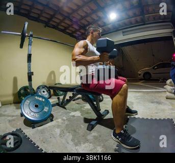Muskulöser Mann in Tanktop und roten Shorts, während er mit Kurzhanteln auf einer Hantelbank sitzt. Stockfoto
