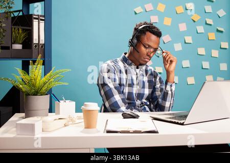 Müder Unternehmensberater am Schreibtisch mit seinem Laptop, mit Headset und Brille. Erschöpfter Mitarbeiter, der tief in Gedanken steckt, an einer Herausforderung für ein Startup-Projekt arbeitet und versucht, Lösungen zu finden. Stockfoto