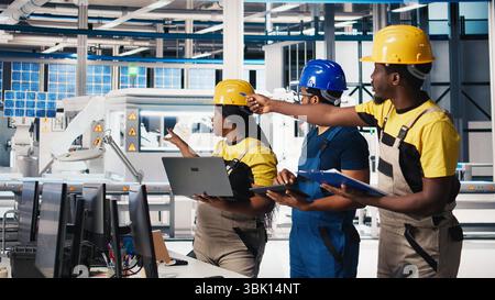 Gruppe schwarzer Mitarbeiter im Werk für Solarpaneele, die Daten auf Geräten und Dokumenten überprüfen. Ingenieure von Photovoltaik-Anlagen, die an innovativen Solarzellen arbeiten, entwickeln einen Prototypen. Kamera B.. Stockfoto