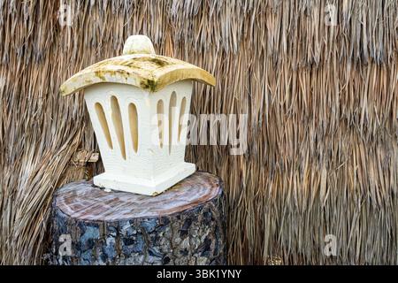Die weiße Laterne sitzt auf einem Baumstamm. Die Laterne besteht aus Beton und hat eine braune Oberseite. Der Baumstamm ist mit trockenen Blättern und Zweigen bedeckt Stockfoto
