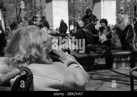 Washington Square Park, eine ältere Frau sieht sich eine Straßenaufführung an. Dieser Moment fängt das Wesen der Gemeinschaft und der generationenübergreifenden Verbindung ein. Stockfoto