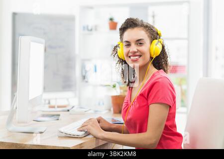 Frau tippt auf weißer Tastatur am Schreibtisch im Heimbüro mit Monitor und gelben Kopfhörern Stockfoto