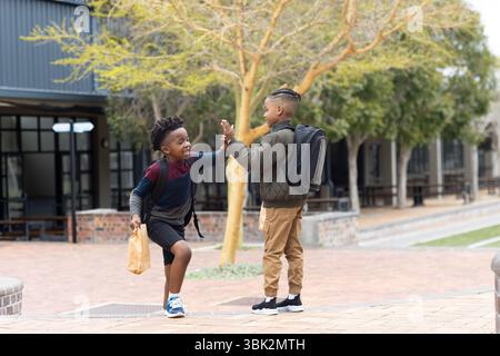 Verschiedene Jungs, die außerhalb der Schule High-Five sind, Rucksäcke tragen und lächeln, Freundschaft genießen Stockfoto