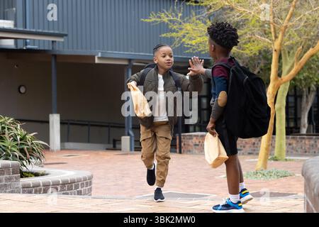 Verschiedene Schulfreunde sind draußen High-Five, tragen Lunchtaschen und tragen Rucksäcke Stockfoto
