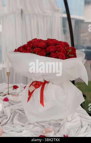 Ein atemberaubender Blick auf einen großen Blumenstrauß aus leuchtenden roten Rosen, elegant präsentiert in knusprigem weißem Geschenkpapier, auf einem Bett mit verstreuten Blütenblättern Stockfoto