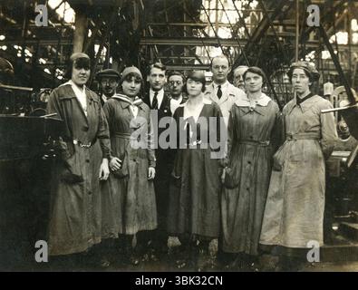 England. Um 1916 – Foto einer Gruppe männlicher und weiblicher Munitionsarbeiter in einer Fabrik während des Ersten Weltkriegs. Die Frauen tragen Overalls, Kopftücher und Hüte. Die zweite von rechts trägt das „On war Service“-Emblem einer Frau am Gürtel. Dieses Emblem wurde ab 1916 nur an Frauen ausgestellt, die in dringenden Kriegsarbeiten tätig waren. Hinter der Gruppe stehen riemengetriebene Maschinen. Stockfoto