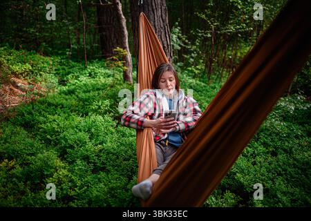 Eine Frau in lässigen Lagen sitzt in einer orangen Hängematte im Wald und schaut auf ihr Telefon im üppigen Grün. Stockfoto