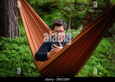 Ein Mann mit Brille und Bart sitzt in einer orangen Hängematte im Wald und konzentriert sich während eines friedlichen Moments draußen auf sein Smartphone. Stockfoto