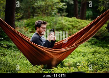 Ein Mann entspannt sich in einer orangen Hängematte im Wald, hält sein Handy mit beiden Händen und lächelt, während er von üppigem Grün umgeben ist. Stockfoto